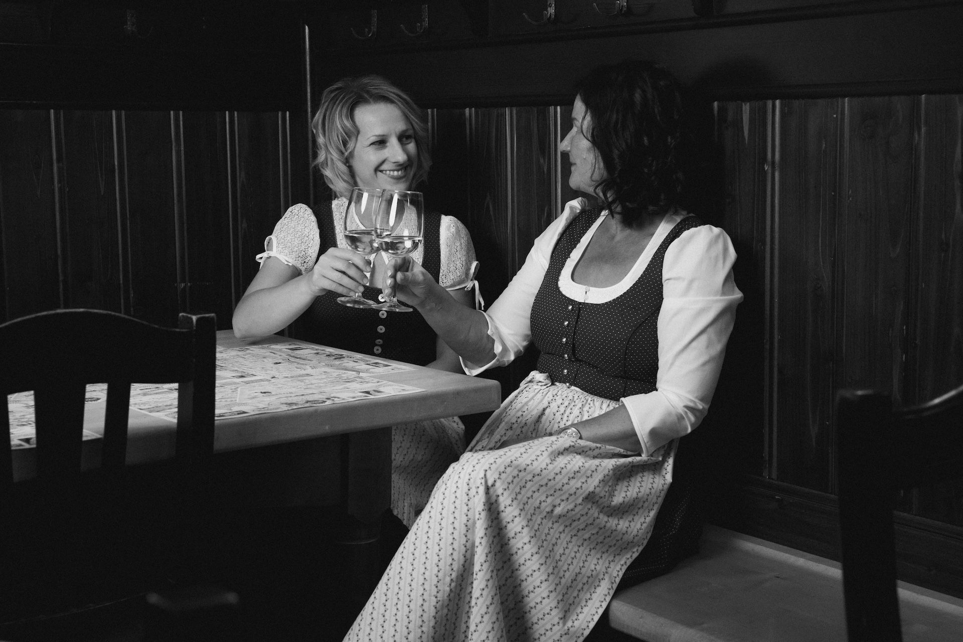 Zwei Frauen in Tracht sitzen an einem Tisch und stoßen mit einem Glas Wein an